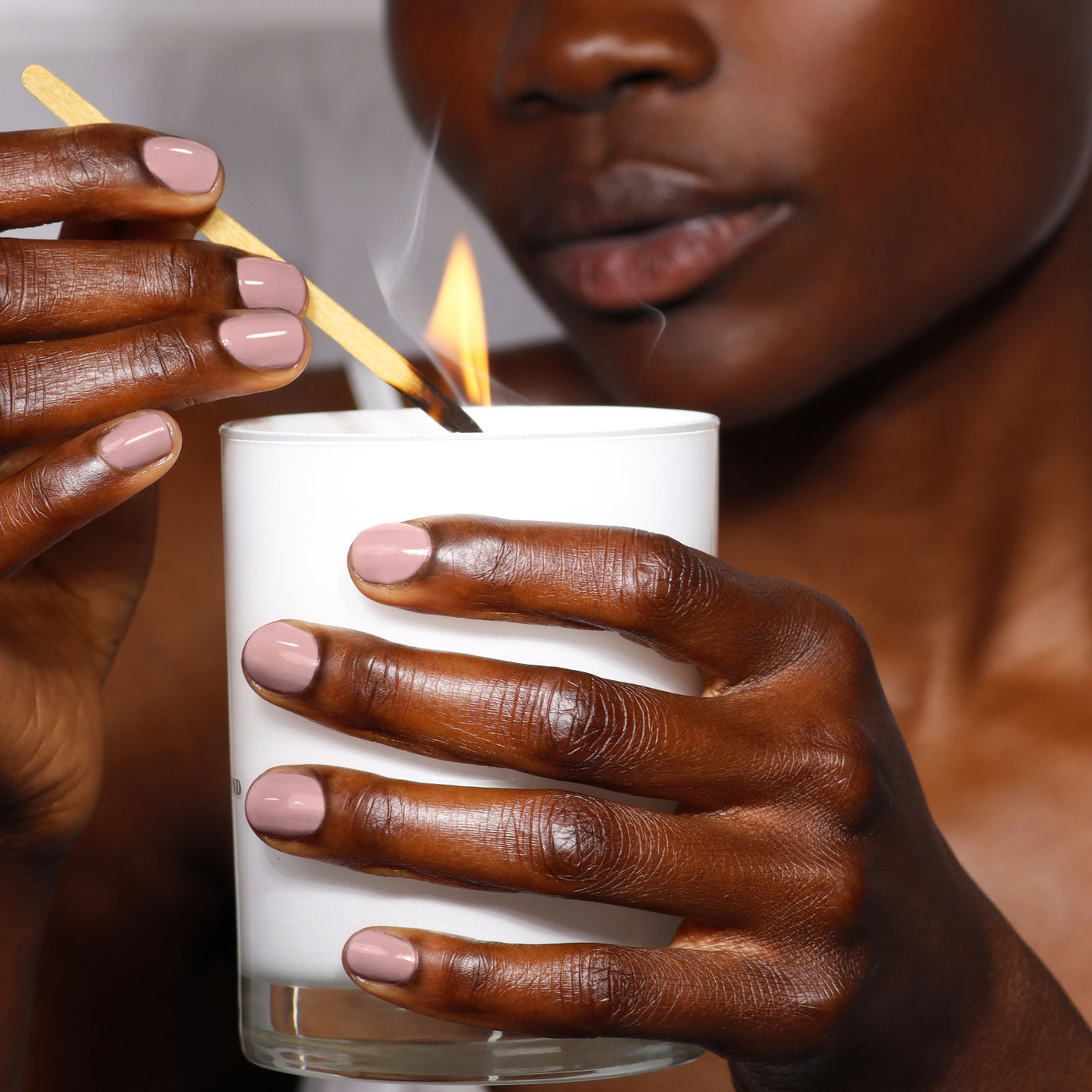 A person lights a white candle with a matchstick, highlighting hands painted in LONDONTOWN's "Do Not Disturb" nail polish, featuring neutral hues with subtle mauve undertones.