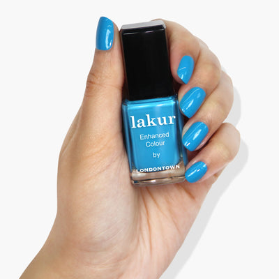 A hand with freshly painted neon blue nails holds a bottle of "Cabana Boy (Professional)" nail polish by LONDONTOWN in matching blue shade.