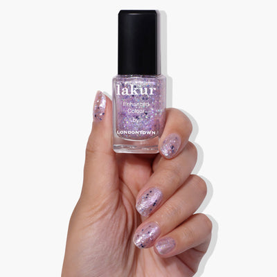 A hand holding a bottle of LONDONTOWN's Grape Fizz (Professional) nail polish. The nails are painted with a delightful holographic purple glitter polish that perfectly matches the bottle.